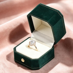 Könnte beinhalten: Ein goldener Verlobungsring mit einem großen, runden, klaren Edelstein wird in einer offenen, dunkelgrünen Samt-Ringbox präsentiert. Die Box hat eine weiße Innenseite und einen goldenen Verschluss. Die Ringbox befindet sich auf einer rosa Samtoberfläche.