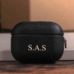 May include: Black leather AirPod case with the initials "S.A.S" in gold lettering. The case has a textured surface and a silver-colored zipper. The case is sitting on a white surface.