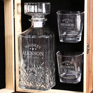 May include: A clear glass whiskey decanter and two matching glasses are displayed in a wooden box. The decanter and glasses are etched with the words "Whiskey Henson" and "Est. 2025". The decanter has a geometric design.