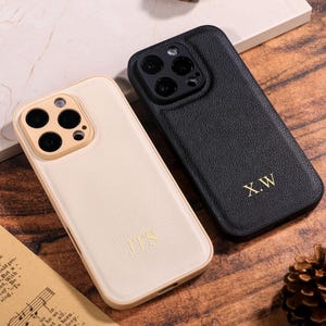 May include: Two leather phone cases, one in cream and one in black. The cream case features the gold initials "JTS", and the black case has "X.W". Both cases have a camera aperture and a smooth finish.