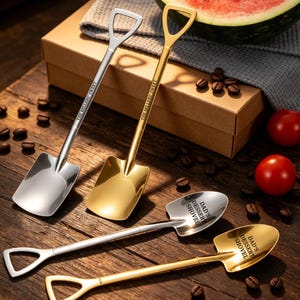 May include: Four metal spoons shaped like shovels, including two with square heads and two with rounded heads. The silver and gold spoons feature engraved text reading THE MILLERS' SWEET or DAD'S DESSERT SHOVEL on the handles or bowls of the spoons.
