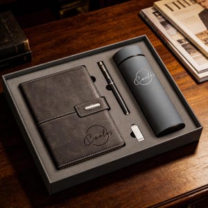 Puede incluir: Un juego de regalo en una caja gris, con un cuaderno de cuero gris oscuro con un cierre, un bolígrafo negro y un vaso térmico gris. El cuaderno y el vaso están personalizados con el nombre "Carlos".