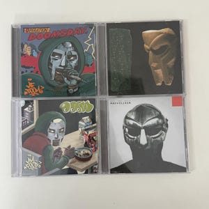 May include: Four CD cases featuring album art. The top left case reads "Operation Doomsday" with a green-hooded figure. The top right case has a gold mask. The bottom left case reads "MM FOOD" and the bottom right case reads "Madvillain".