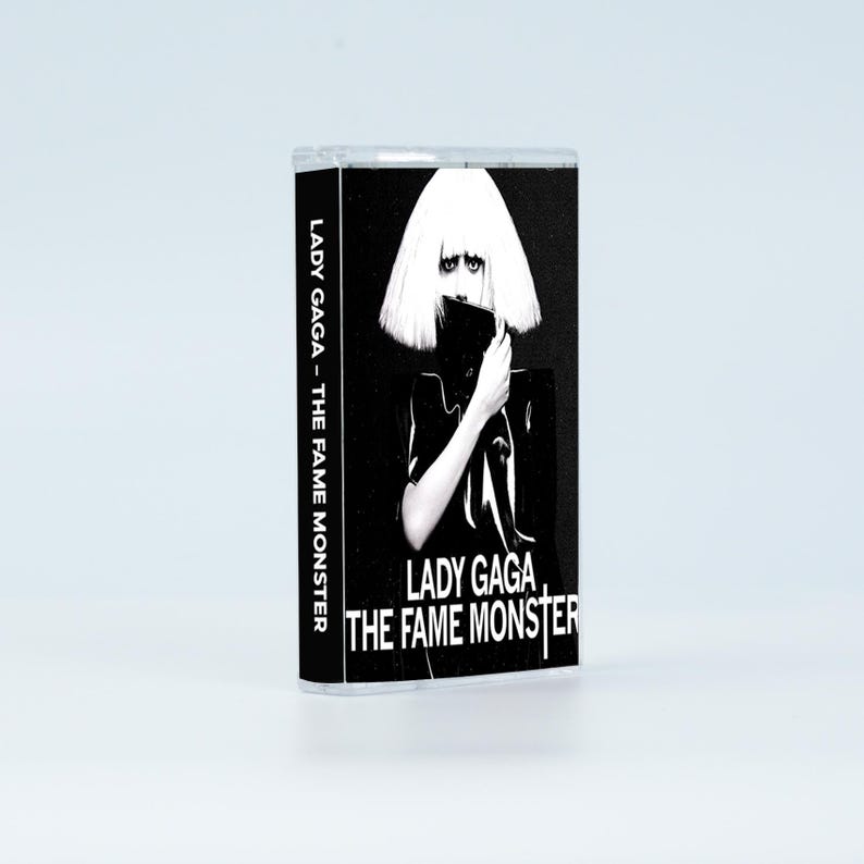 May include: A cassette tape case featuring the album cover for Lady Gaga - The Fame Monster. The black and white artwork displays a person with a blonde bob hairstyle, holding a black object near their face. The text reads Lady Gaga - The Fame Monster.