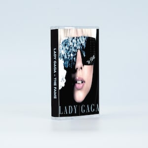 May include: A cassette tape of Lady Gaga's album The Fame. The cover features a close-up of a face wearing dark, crystal-embellished sunglasses. The text LADY GAGA - THE FAME is printed vertically on the black spine and horizontally at the bottom.