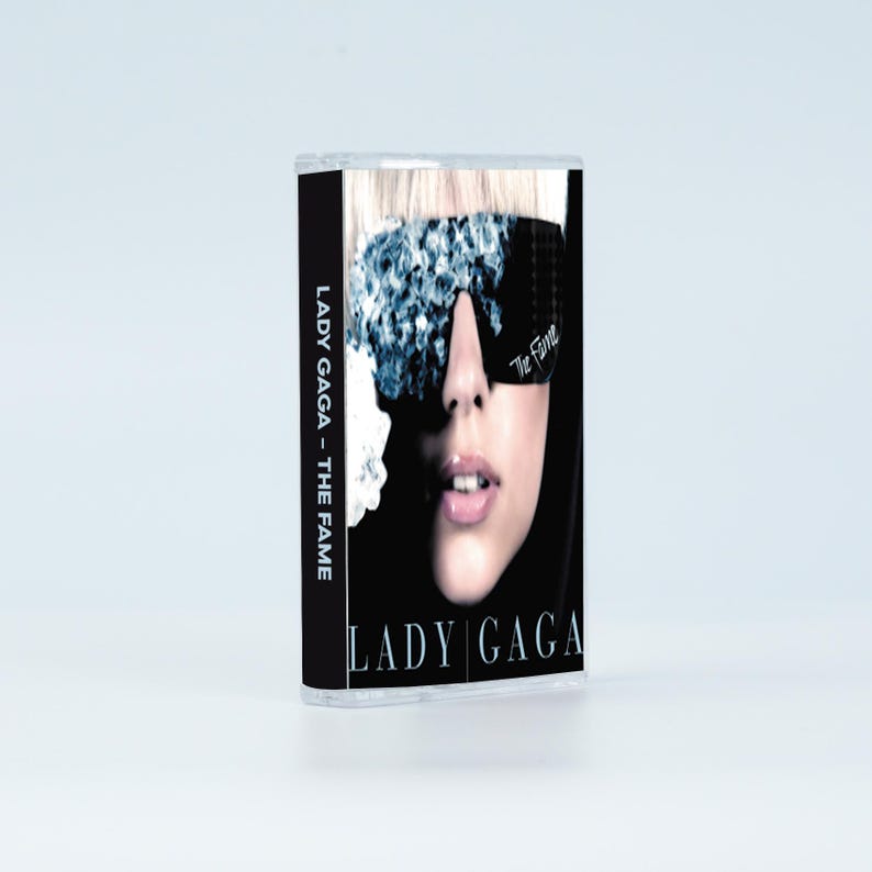 May include: A cassette tape of Lady Gaga's album The Fame. The cover features a close-up of a face wearing dark, crystal-embellished sunglasses. The text LADY GAGA - THE FAME is printed vertically on the black spine and horizontally at the bottom.