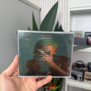 May include: A CD case with a green and brown cover, featuring a tracklist including "NIKES," "IVY," and "PINK + WHITE." The image shows a person holding the CD, with a reflection visible on the case.
