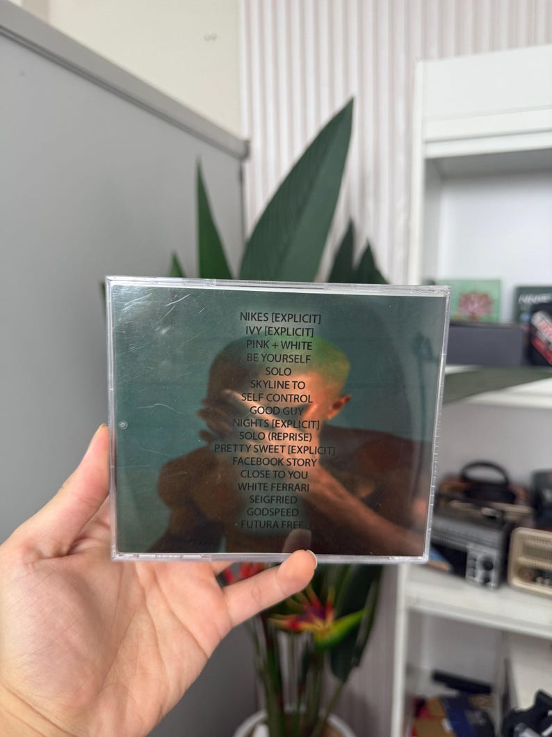 May include: A CD case with a green and brown cover, featuring a tracklist including "NIKES," "IVY," and "PINK + WHITE." The image shows a person holding the CD, with a reflection visible on the case.