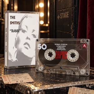 May include: A clear plastic cassette tape with black magnetic tape and white text reading 50 and ZAPAS, alongside a cassette case featuring a black and white portrait and the text THE SMITHS RANK, plus a vintage Odeon Theatre ticket stub.