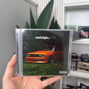 May include: A CD case featuring the album "nostalgia, ULTRA." The cover art displays an orange car parked in a grassy area with trees. The text "nostalgia, ULTRA." is in white font. A parental advisory warning is visible.
