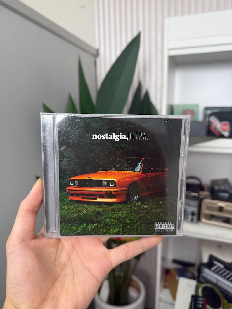 May include: A CD case featuring the album "nostalgia, ULTRA." The cover art displays an orange car parked in a grassy area with trees. The text "nostalgia, ULTRA." is in white font. A parental advisory warning is visible.