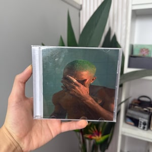 May include: A CD case featuring album art of a person with green hair covering their face. The artwork has a teal background. The CD case is held in a hand.