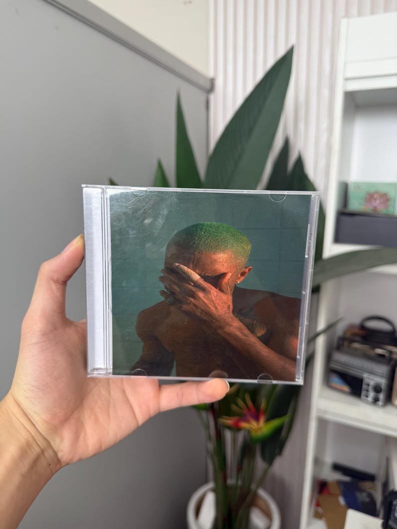 May include: A CD case featuring album art of a person with green hair covering their face. The artwork has a teal background. The CD case is held in a hand.
