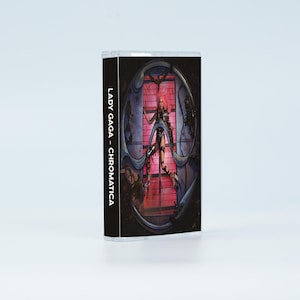 May include: A cassette tape case featuring the album cover for Lady Gaga - Chromatica. The cover art displays a figure in a dark, metallic, circular structure against a bright pink grid background. The spine text reads LADY GAGA - CHROMATICA.