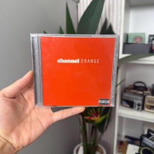 May include: A CD case with a bright orange cover. The text "channel ORANGE" is printed in white. The case has an advisory warning sticker. The CD is held in a hand.