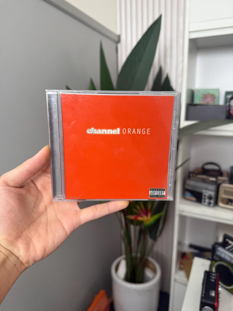 May include: A CD case with a bright orange cover. The text "channel ORANGE" is printed in white. The case has an advisory warning sticker. The CD is held in a hand.