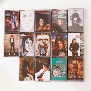 May include: A collection of vintage Michael Jackson cassette tapes. The album covers feature portraits of the artist and album titles like "History," "Thriller," and "Ben." The tapes are arranged on a white surface.