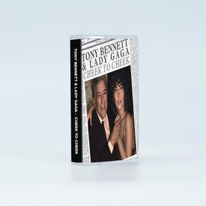 May include: A cassette tape case featuring the album cover for Tony Bennett & Lady Gaga's Cheek to Cheek. The cover design mimics a newspaper layout with the artists' names and album title in bold black text above a photograph of the two musicians.