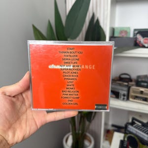 May include: A CD case with an orange insert featuring a track list. The text includes song titles such as "Start", "Sweet Life", and "Golden Girl". The CD is held in a person's hand.
