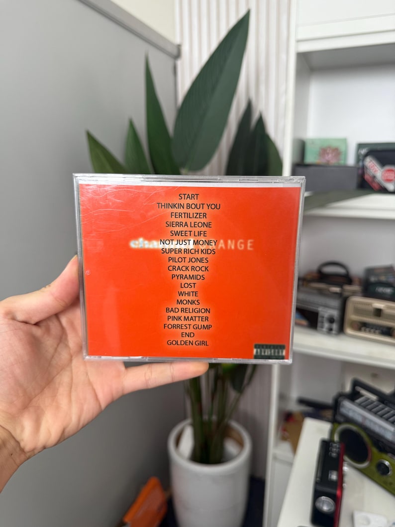 May include: A CD case with an orange insert featuring a track list. The text includes song titles such as "Start", "Sweet Life", and "Golden Girl". The CD is held in a person's hand.