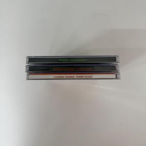 May include: Three compact disc cases stacked on top of each other. The top case is labeled "BLONDE - FRANK OCEAN" in white text on a black background. The middle case reads "NOSTALGIA, ULTRA - FRANK OCEAN". The bottom case is labeled "CHANNEL ORANGE - FRANK OCEAN".