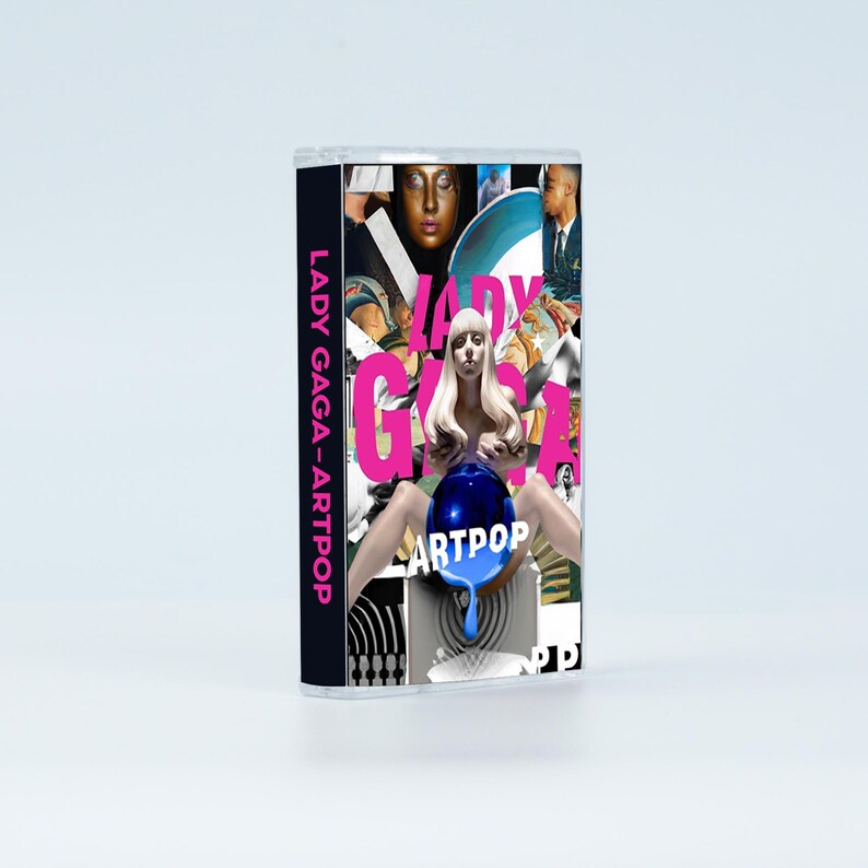 May include: A cassette tape case featuring the Lady Gaga Artpop album cover. The artwork displays a collage of images including a central figure with blonde hair holding a blue sphere, with the text LADY GAGA and ARTPOP printed in pink and white.