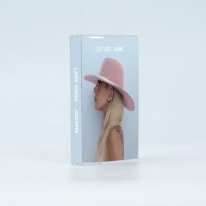 May include: A clear plastic cassette case featuring the album cover for Lady Gaga's Joanne. The cover art displays a side profile of a person with long blonde hair wearing a wide-brimmed light pink hat against a pale blue background.
