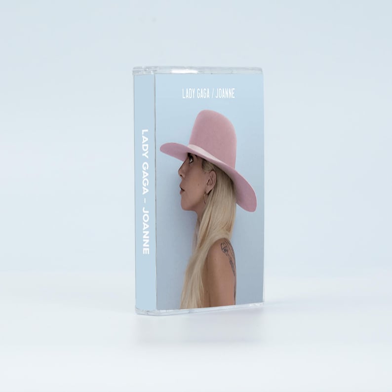 May include: A clear plastic cassette case featuring the album cover for Lady Gaga's Joanne. The cover art displays a side profile of a person with long blonde hair wearing a wide-brimmed light pink hat against a pale blue background.