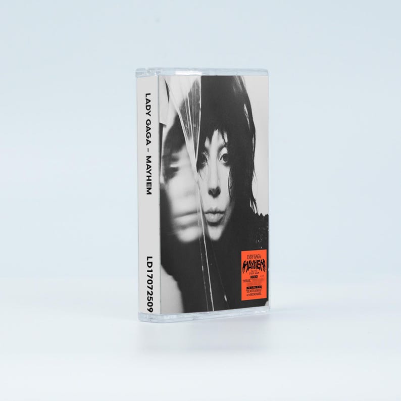 May include: A cassette tape case featuring a black and white portrait of Lady Gaga on the cover. The spine displays the text LADY GAGA - MAYHEM and the code LD17072509. A small orange rectangular sticker with black text is on the lower right corner.