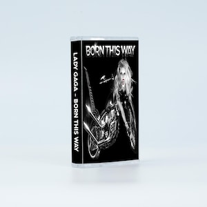 May include: A cassette tape featuring the album Born This Way by Lady Gaga. The cover art displays a black and white image of a person with long hair merged with a motorcycle. The text Born This Way appears at the top, and Lady Gaga - Born This Way is printed vertically on the side spine of the clear plastic case.