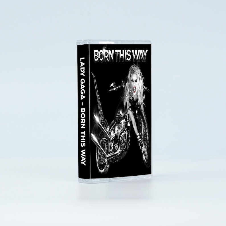 May include: A cassette tape featuring the album Born This Way by Lady Gaga. The cover art displays a black and white image of a person with long hair merged with a motorcycle. The text Born This Way appears at the top, and Lady Gaga - Born This Way is printed vertically on the side spine of the clear plastic case.