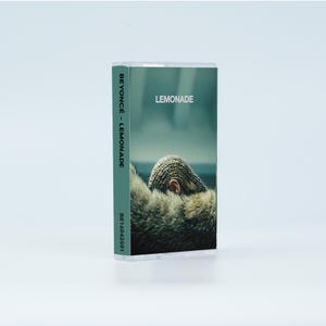 May include: A cassette tape case featuring the album Lemonade by Beyonc&eacute;. The cover art displays a person with braided hair wearing a fur garment. The spine includes the text BEYONC&Eacute; - LEMONADE and the code BE16042501 in white lettering.