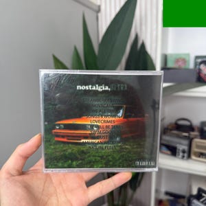 May include: A CD case with the album title "nostalgia, ULTRA" and a tracklist including "Strawberry Swing" and "Novacane." The cover art features an orange car in a forest setting. The CD is held in a person's hand.