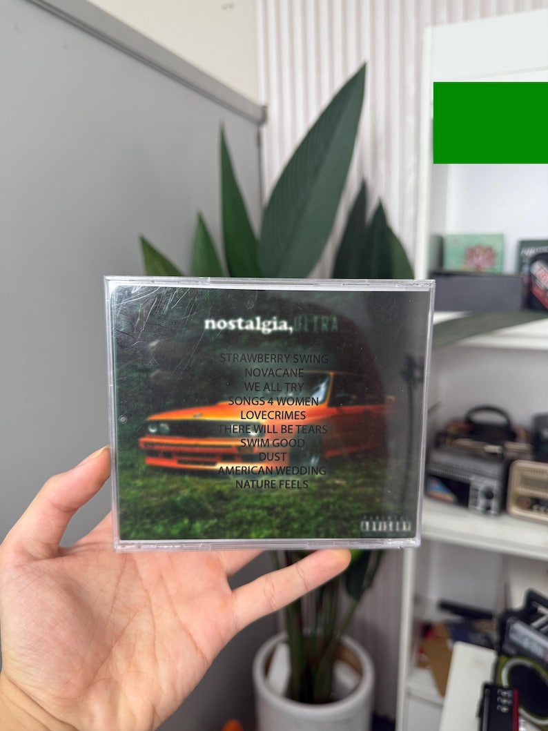 May include: A CD case with the album title "nostalgia, ULTRA" and a tracklist including "Strawberry Swing" and "Novacane." The cover art features an orange car in a forest setting. The CD is held in a person's hand.