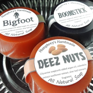 May include: Three round bars of handmade soap with labels. The labels are white with black text and feature a different design and name for each soap. The soaps are orange, black, and brown. The labels read "Bigfoot", "Boomstick", and "Deez Nuts".
