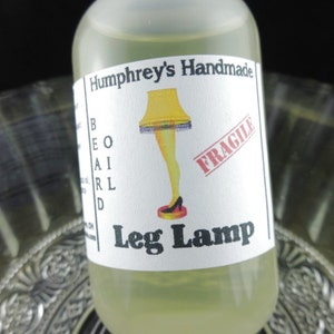 May include: A clear glass bottle with a white label featuring a cartoon leg lamp design. The label reads "Humphrey's Handmade" and "Leg Lamp" in black text. The bottle contains a light yellow liquid.