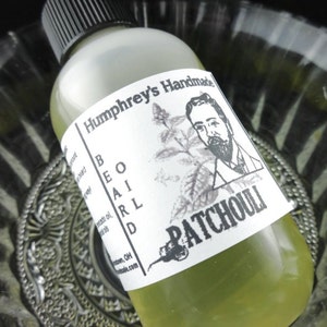 May include: A clear glass bottle with a black cap and a white label. The label says "Humphrey's Handmade Beard Oil Patchouli". The bottle is sitting on a silver and white patterned surface.