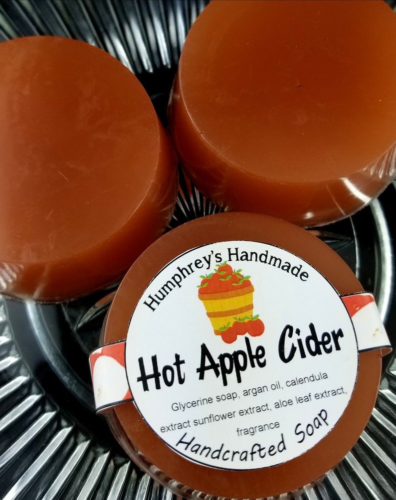 May include: Three round bars of brown soap with a white label that reads "Humphrey's Handmade Hot Apple Cider Glycerine soap, argan oil, calendula extract sunflower extract, aloe leaf extract, fragrance Handcrafted Soap".