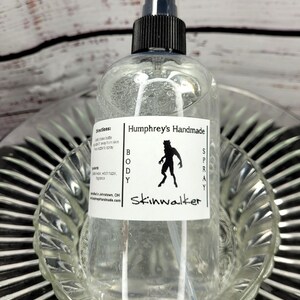 May include: Clear body spray bottle with a black and white label that reads "Humphrey's Handmade BODY SPRAY Skinwalker." The label features a silhouette of a creature. The bottle has a black spray nozzle and is sitting in a clear glass dish.