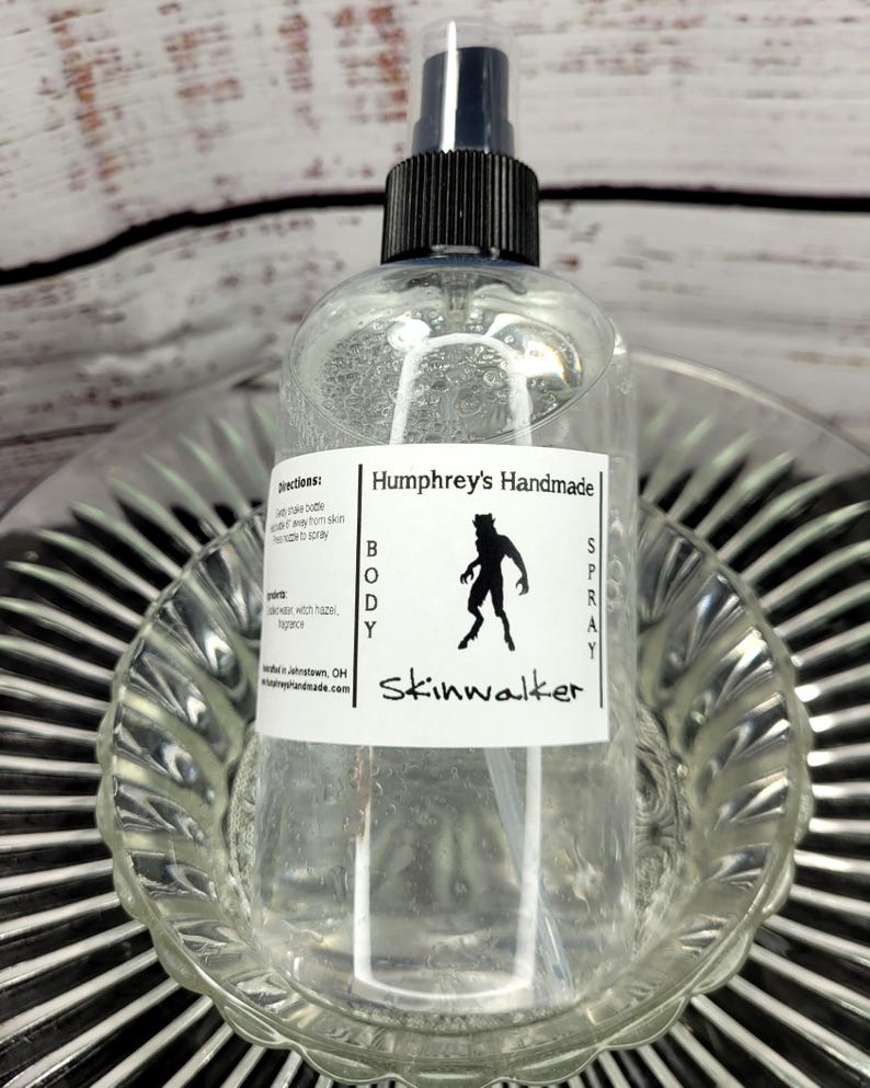 May include: Clear body spray bottle with a black and white label that reads "Humphrey's Handmade BODY SPRAY Skinwalker." The label features a silhouette of a creature. The bottle has a black spray nozzle and is sitting in a clear glass dish.
