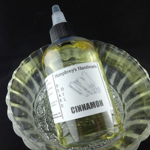 May include: A clear glass bottle with a black cap and a white label that says "Humphrey's Handmade Beard Oil Cinnamon". The bottle is sitting in a clear glass bowl.