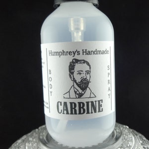 May include: A clear glass bottle with a black spray top and a white label. The label says "Humphrey's Handmade Body Spray" and "CARBINE" in black text. A black and white illustration of a man with a beard is on the label.