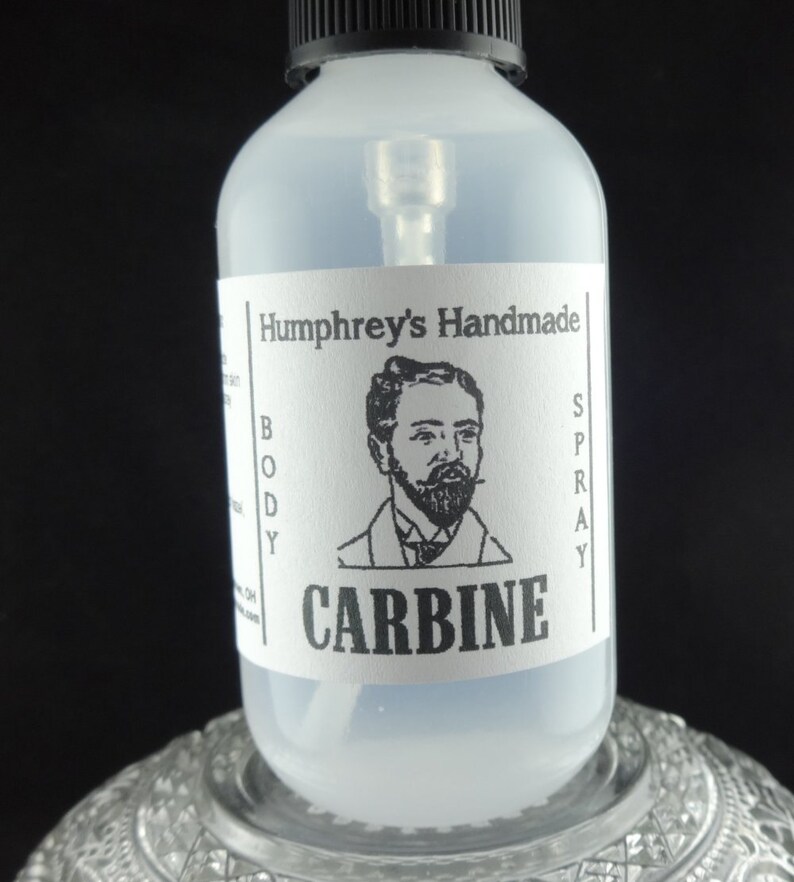 May include: A clear glass bottle with a black spray top and a white label. The label says "Humphrey's Handmade Body Spray" and "CARBINE" in black text. A black and white illustration of a man with a beard is on the label.
