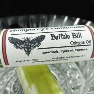 May include: A small, clear glass bottle with a white label that reads "Humphrey's Handmade Buffalo Bill Cologne Oil". The label also includes a black and white illustration of a moth, the ingredients, and the text "net wt. 33 oz/10 ml".