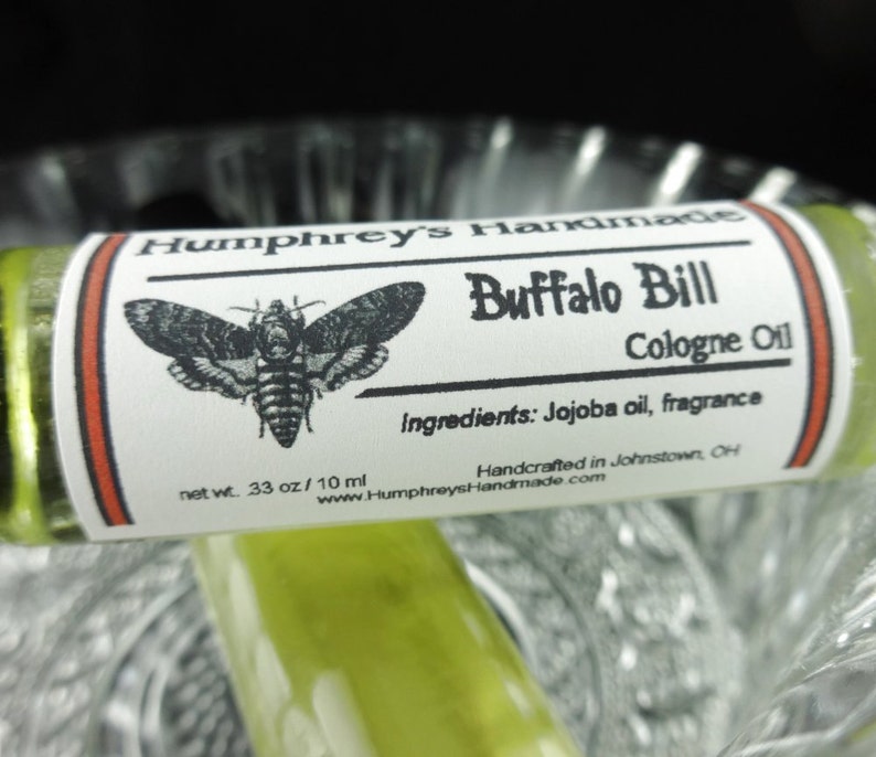 May include: A small, clear glass bottle with a white label that reads "Humphrey's Handmade Buffalo Bill Cologne Oil". The label also includes a black and white illustration of a moth, the ingredients, and the text "net wt. 33 oz/10 ml".
