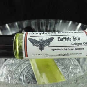 May include: A glass roll-on bottle with a white label that says "Humphrey's Handmade Buffalo Bill Cologne Oil" with a black and white illustration of a moth. The label also says "Ingredients: Jojoba oil, fragrance" and "Handcrafted in Johnstown, OH".