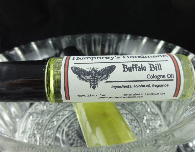 May include: A glass roll-on bottle with a white label that says "Humphrey's Handmade Buffalo Bill Cologne Oil" with a black and white illustration of a moth. The label also says "Ingredients: Jojoba oil, fragrance" and "Handcrafted in Johnstown, OH".