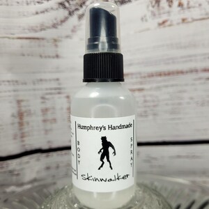 May include: A clear glass spray bottle with a black pump and cap. The label reads "Humphrey's Handmade BODY SPRAY Skinwalker" with a silhouette of a werewolf. The bottle is sitting on a decorative glass dish.