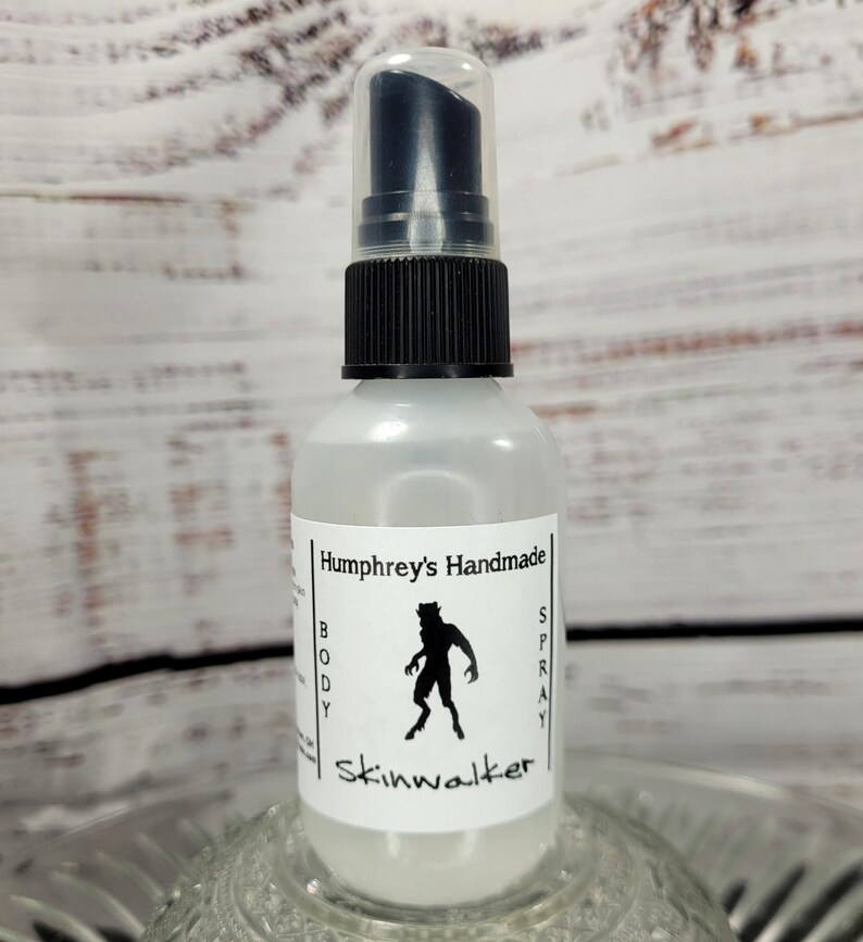 May include: A clear glass spray bottle with a black pump and cap. The label reads "Humphrey's Handmade BODY SPRAY Skinwalker" with a silhouette of a werewolf. The bottle is sitting on a decorative glass dish.