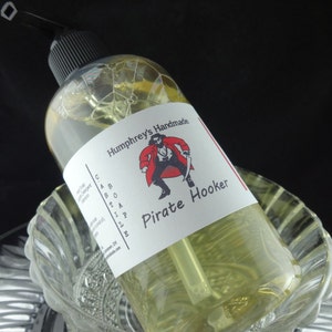 May include: A clear glass bottle with a pump dispenser filled with a yellow liquid. The bottle has a white label with red and black illustrations and the text "Humphrey's Handmade Castile Soap Pirate Hooker".